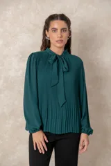 Blusa plisada de color verde oscuro, con mangas largas terminadas en puños elásticos y cuello alto con lazo frontal para anudar.