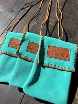 Bandolera compacta de gamuza turquesa con solapa, tira fina de cuero para colgar al hombro o cruzada, costuras artesanales visibles en los bordes y parche frontal de cuero con el logo grabado de SierraMora.