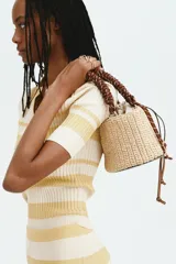 Cartera tipo tote en paja sintética trenzada, con estructura cilíndrica. Cuenta con un bolso interior de lona color beige con cordón ajustable, asa superior trenzada de cuerda marrón y detalles en ecuero.