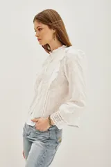 Camisa blanca de algodón con diseño romántico, cuello alto con volados, cierre frontal de botones y detalles de encaje y bordados calados. Presenta mangas largas con puños de encaje y volados frontales.