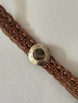 Cinto trenzado de cuero color camel con hebilla circular metálica grabada y detalle de piedra central.