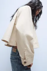 Trench corta de color beige, con cuello solapa, manga larga y cierre frontal con botones oscuros. Presenta bolsillos de vivo en el delantero y un diseño de corte recto.
