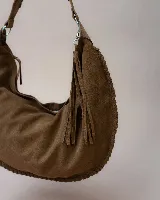 Bolso tipo hobo de gran tamaño, confeccionado en gamuza color marrón oscuro. Presenta un diseño artesanal con costuras visibles y detalles de tientos en el cuerpo, asa integrada con pespuntes a mano y cierre superior.