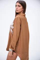 Camisa marrón de manga larga con puños anchos, cuello camisero y botones frontales. Presenta bordados florales en blanco con lentejuelas y mostacillas en la parte delantera.