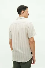 Camisa de manga corta confeccionada en una mezcla de lino y algodón, con diseño a rayas verticales en tonos claros. Presenta cuello camisero con botones, cierre frontal abotonado y un bolsillo en el pecho con logo bordado.