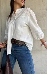 Camisa blanca de estilo romántico con cuello mao, botones frontales y mangas tres cuartos abullonadas con detalle de frunces. Presenta un diseño con textura sutil y costuras decorativas en la parte superior delantera.