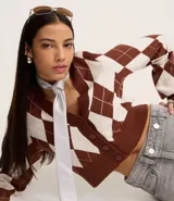 Cardigan cropped de tejido jacquard con diseño de rombos argyle en color marrón y blanco. Presenta escote en V, manga larga y cierre frontal con botones marrones.