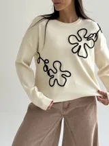 Sweater de punto color crema con cuello redondo, mangas largas y puños acanalados. Presenta un diseño floral bordado con cordón negro en el frente.