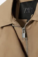 Chaqueta tipo bomber de color beige, con cuello solapa y aplicación extraíble de pelo sintético. Presenta cierre frontal con botones a presión ocultos, bolsillos delanteros y bajo con elástico.