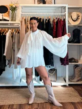 Blusa blanca de corte holgado y diseño plisado, con cuello alto y lazo para anudar al frente. Presenta mangas largas amplias con puños ajustados y una caída fluida y elegante.