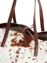 Bolso tipo tote de piel de vaca, con estampado moteado en blanco y marrón. Presenta asas de cuero marrón oscuro y costuras decorativas en forma de X en el centro frontal.