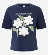 Remera de algodón color blanco, con cuello redondo y manga corta. Presenta un estampado con la frase 'Soy flor. Soy latina' y flores bordadas en relieve con textura tejida.
