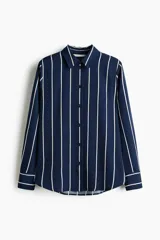 Camisa de manga larga con cuello clásico, cierre frontal abotonado y puños con botón. Presenta un diseño de rayas verticales blancas sobre fondo azul marino, con bajo redondeado.