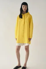 Cazadora impermeable de acabado engomado en color amarillo, con capucha ajustable, cierre frontal con botones a presión, bolsillos laterales y mangas largas ajustables.