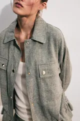 Chaqueta corta gris de tejido texturizado con cuello solapa, manga larga con trabilla, bolsillos delanteros con solapa y cierre frontal de botones metálicos.