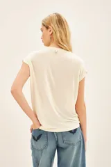 Remera de viscosa color crema, de calce suelto, escote redondo y manga corta. Presenta una estampa frontal centrada con el dibujo de un rostro y manos, y un bordado de un ojo con pedrería.