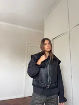 Chaleco de cuero sintético negro con interior de corderito, cierre frontal metálico y cinturón ajustable en la cintura.