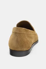 Mocasín plano de mujer en serraje color camel, con antifaz y copete. Presenta punta redonda y suela baja de goma negra.
