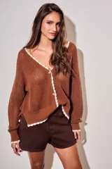 Cardigan de punto color marrón, de corte holgado y manga larga, con escote en V y cierre frontal de botones. Presenta detalles de terminación festoneada en color blanco en los bordes.