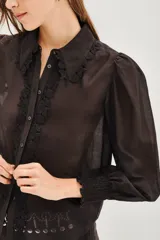 Camisa negra de manga larga con diseño de hombros abullonados y puños elásticos. Presenta un cuello clásico y una tapeta frontal con botones, decorada con detalles de encaje floral a lo largo de la abertura.
