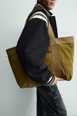 Bolso tipo shopper de gran capacidad, confeccionado en tejido con textura de pana. Cuenta con dos asas de mano resistentes, un bolsillo frontal plano y cierre principal mediante botón a presión. El interior incluye un bolsillo con cierre de cremallera.