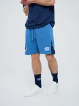 Bermuda deportiva Umbro del Club Nacional de Football, modelo Nexo para hombre, en color azul marino con detalles en rojo y blanco. Cuenta con el escudo del club bordado en la pierna derecha y el logo de Umbro en la izquierda, además de cordón ajustable en la cintura.