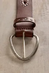 Cinturón de cuero marrón con calados en forma de gota y tachas metálicas a lo largo de la tira. Cuenta con hebilla metálica ovalada y ajuste regulable.