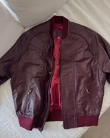 Campera tipo bomber de cuero ovino color marrón oscuro, con cuello de tejido acanalado y cierre frontal con cremallera.