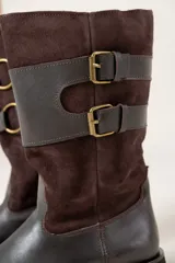 Bota de caña media confeccionada en una combinación de cuero liso y gamuza color chocolate. Presenta un diseño con cierre lateral, doble hebilla decorativa en el lateral externo y suela plana con pequeño tacón.