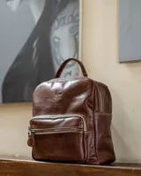 Mochila de cuero natural color negro, con diseño clásico y refinado. Cuenta con un compartimento principal con cierre, un bolsillo frontal con cierre y un asa superior. Presenta el logo de la marca grabado en la parte frontal.