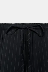 Pantalón de tiro medio, color negro con rayas diplomáticas finas blancas. Cintura elástica ajustable con cordones y bajo abullonado con elástico.
