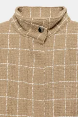Chaqueta bomber de color beige con diseño a cuadros blancos, cuello subido, manga larga con trabillas en los hombros, bolsillos delanteros y bajo con elástico. Cierre frontal con cremallera y botones a presión.