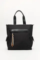 Bolso tipo tote color negro, con doble asa superior y correa desmontable. Presenta un bolsillo frontal con solapa, bolsillos laterales y un detalle colgante decorativo con tiras de colores.