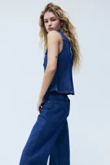 Chaleco de denim azul rígido de algodón, con cuello camisero, cierre frontal de botones metálicos, bolsillos con solapa en el pecho y detalle de peplum en la parte inferior.
