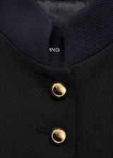Chaqueta estructurada de corte recto en color negro, con cuello mao y cierre frontal de botones dorados con efecto joya. Presenta manga larga con puños abotonados y forro interior.