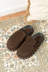 Zuecos de gamuza color marrón chocolate, con base de goma negra, tachas plateadas alrededor de la base y plantilla acolchada.