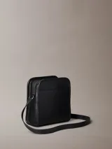 Cartera tipo reporter de Calvin Klein confeccionada en piel sintética suave de color negro. Presenta un diseño cuadrado con monograma tonal grabado en el frente, correa ajustable para cruzar, cierre de cremallera superior y bolsillo exterior trasero.
