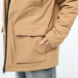 Parka ligera color beige con capucha ajustable, cierre frontal con cremallera y parche decorativo con texto en la manga.