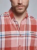 Camisa de franela de manga larga con estampado de cuadros en tonos rojos, rosados y detalles en azul. Presenta cuello con botones, cierre frontal abotonado y un bolsillo en el pecho con detalle de marca.