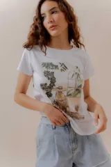 Remera blanca de algodón con cuello redondo y manga corta. Presenta una estampa ilustrada de una mujer con sombrero sentada en un interior, rodeada de plantas y mirando a través de una ventana con vista a palmeras.