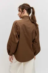 Camisa de manga larga color marrón, con cuello alto con volados, botonadura frontal y detalles calados en las mangas abullonadas.