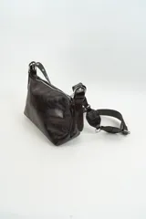 Cartera de hombro color marrón oscuro, confeccionada en cuero sintético con acabado brillante. Presenta una correa larga ajustable con detalles de piezas circulares decorativas y remaches metálicos.