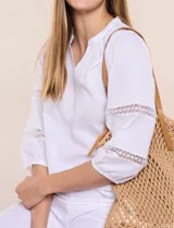 Blusa blanca de calce suelto y caída ligera, con escote en V y cuello Mao pequeño. Presenta mangas tres cuartos abullonadas con elástico en los puños y detalles de calado o puntilla encastrada. Espalda lisa con canesú y leve frunce.