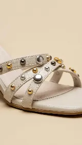 Sandalias de taco ancho de 7 cm, color crema, con tiras cruzadas y cierre de hebilla al tobillo. El diseño presenta múltiples tachas decorativas metálicas en tonos dorados y plateados distribuidas a lo largo de las tiras y en todo el taco.