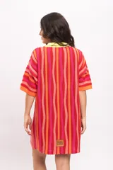 Vestido camisola corto de tejido fresco de algodón, con diseño a rayas verticales en tonos rojos, naranjas y fucsias. Cuenta con cuello polo y botonadura frontal en tono amarillo claro, y mangas cortas holgadas.