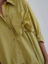 Camisa de popelina color verde oliva, de corte holgado, con cuello clásico, cierre frontal de botones, bolsillo en el pecho con detalle de rosetón a tono y diseño asimétrico más largo en la parte trasera.