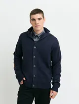 Cardigan de punto color marrón con cuello esmoquin, cierre frontal con botones y presilla superior. Presenta mangas largas con coderas en contraste y bolsillos frontales de ribete.