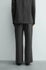 Pantalón de vestir gris oscuro, corte ancho (flare fit) y tiro alto, con bolsillos laterales y falsos bolsillos de vivo en la parte trasera. Presenta un cierre frontal con cremallera y gancho oculto, y botones a presión ocultos en el bajo.