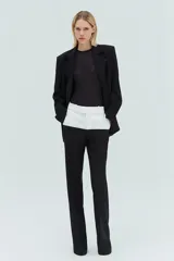 Pantalón de sastrería bicolor, de corte recto y holgado, con cintura alta y trabillas. Cuenta con un diseño en contraste donde la pretina y la parte superior presentan un bloque en color blanco, mientras que el resto de la prenda es negro. Posee bolsillos delanteros y traseros, y cierre frontal con cremallera, botón y corchetes.
