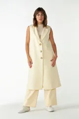 Chaleco largo sin mangas, color crudo/beige claro, con solapas de traje y cierre frontal de tres botones dorados. Presenta bolsillos ojal a la altura de la cadera.